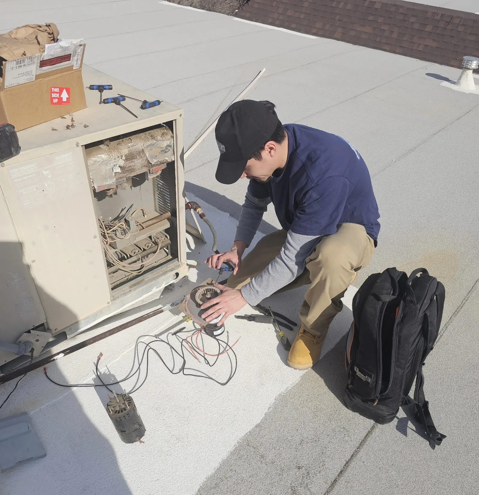 Constant Air Balancing technician in a black cap crouched on a roof beside an open unit, tools and a bag spread on the roof